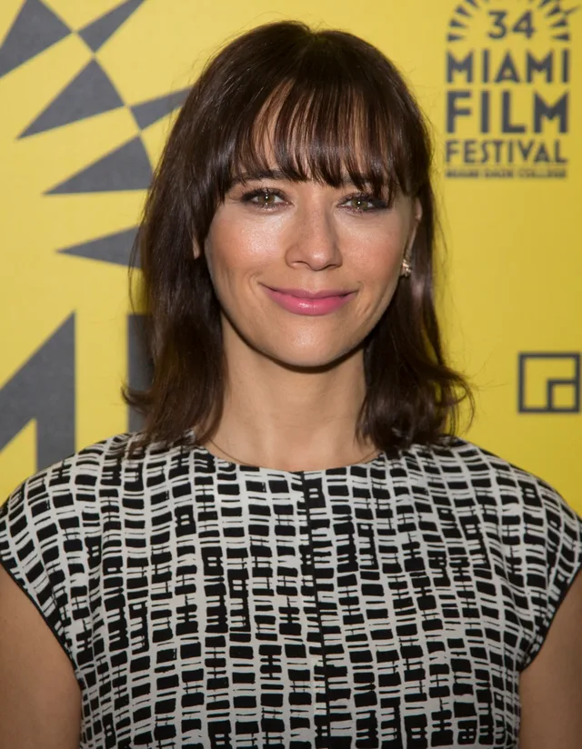 Colour photograph of a smiling woman with a fringe and shoulder-length dark hair, in a black and white patterned top, in front of a yellow festival backdrop.