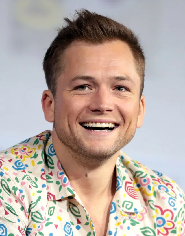 Close photograph of a smiling man with short brown hair in a cream shirt printed with colourful flowers and swirls.
