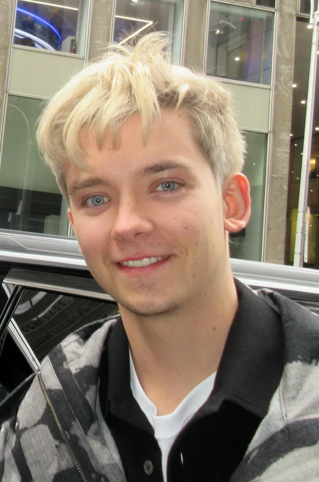 Close photograph of a smiling young man with tousled blond hair and pale blue eyes, in a grey and black patterned jacket, standing outside beside a car.