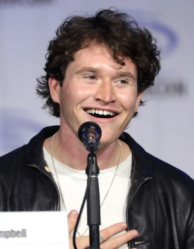 Rohan Campbell smiling at a panel microphone, wearing a black leather jacket over a white t-shirt.