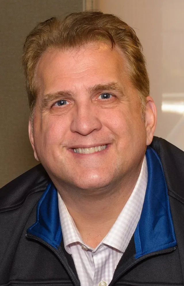 Close portrait of Daniel Roebuck smiling, with short light brown hair, wearing a dark zip jacket with blue trim over a pale checked shirt.