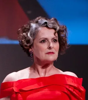 Woman with short curled grey and brown hair in an off-the-shoulder red gown, photographed on a stage.