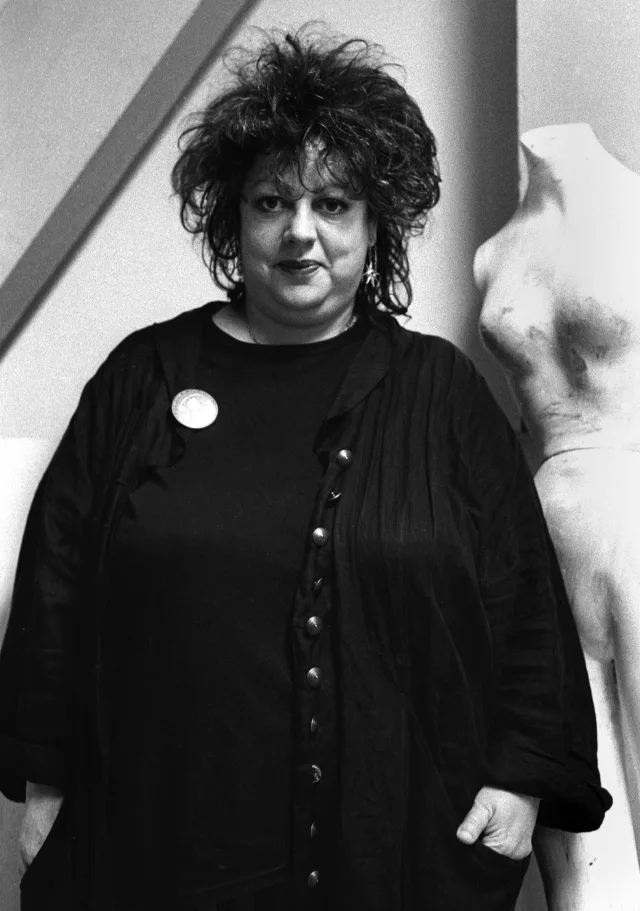 Black and white photograph of a woman with dark, tousled hair in a loose black cardigan with a round badge, standing with hands in pockets.