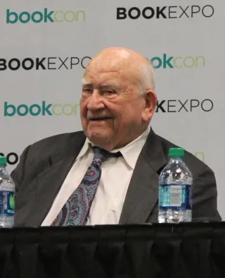 Elderly man in a grey jacket and patterned tie seated at a table at a book convention, looking off to one side.