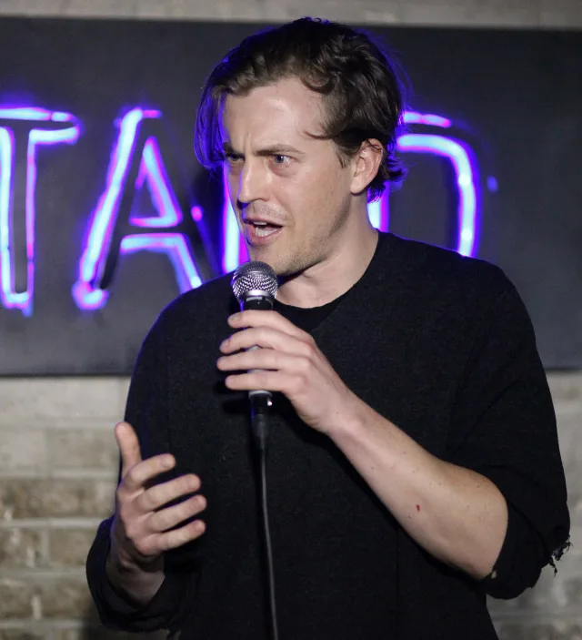 A man in a dark sweater holding a microphone and gesturing as he speaks on stage, with a neon sign and brick wall behind him.