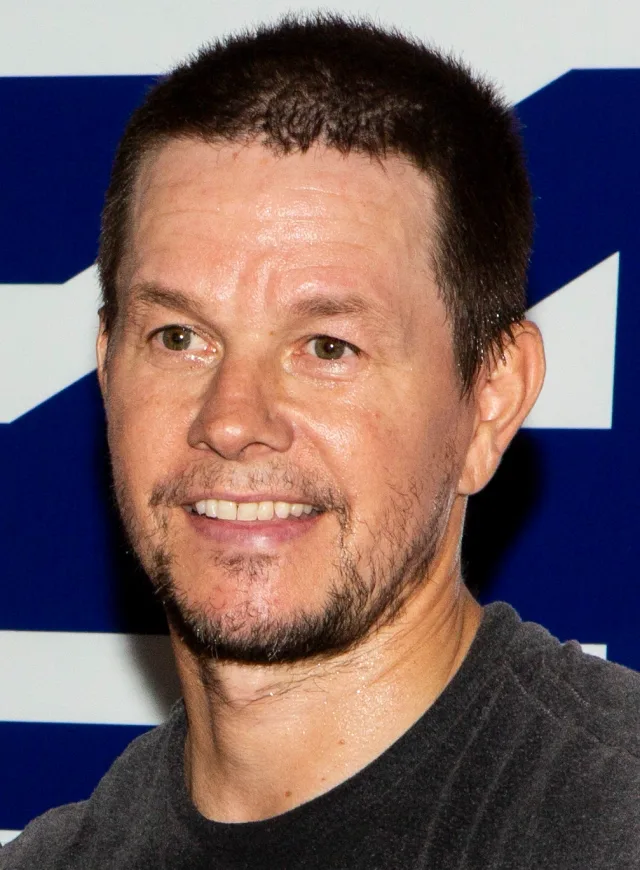 Close-up photograph of a smiling man with short dark hair and stubble, wearing a grey t-shirt in front of a blue and white backdrop.