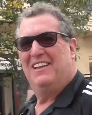 Close-up of a smiling older man with grey hair and dark sunglasses, wearing a black polo shirt outdoors.