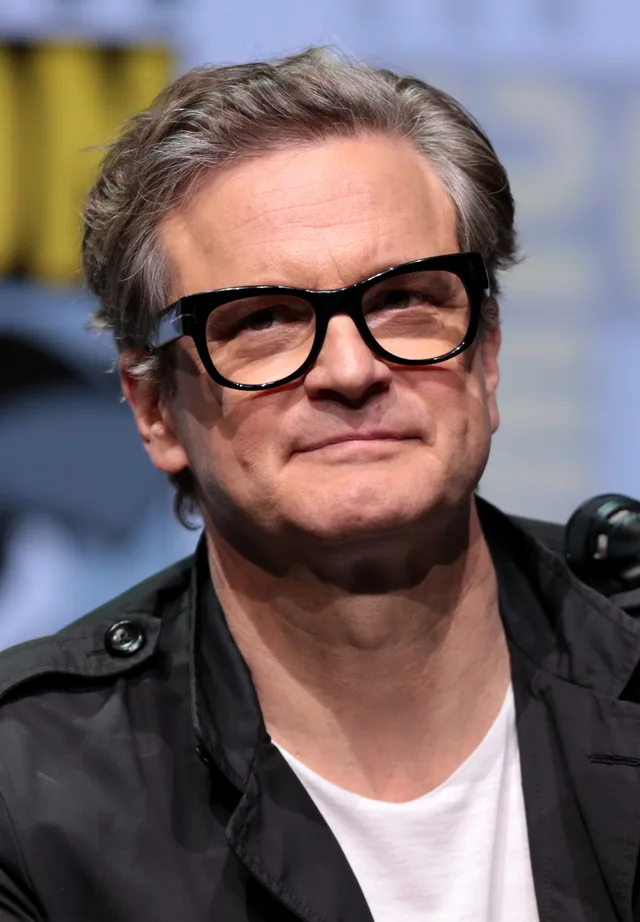 Colin Firth on a convention panel stage, speaking and looking off to one side.