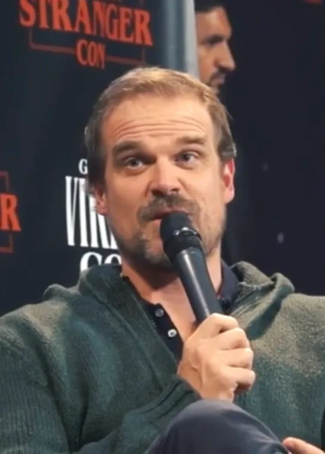 Close photograph of a man with short fair hair and a goatee speaking into a handheld microphone at a fan convention panel, wearing a green zip-neck jumper.