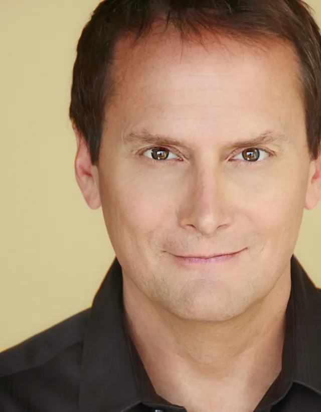 A close head shot of a man with short brown hair and a faint smile, wearing a dark collared shirt against a pale yellow wall.
