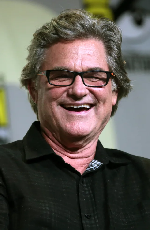 Close-up photograph of a smiling older man with swept grey hair and black-framed glasses, wearing a dark collared shirt.