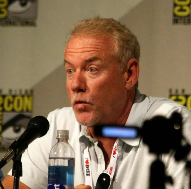 Man with short fair hair and light stubble speaking at a convention panel table, with a microphone and a water bottle in front of him.