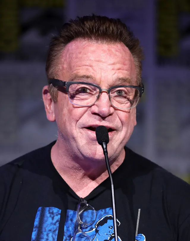 Tom Arnold speaking into a microphone, wearing dark-framed glasses and a black t-shirt with a blue print.