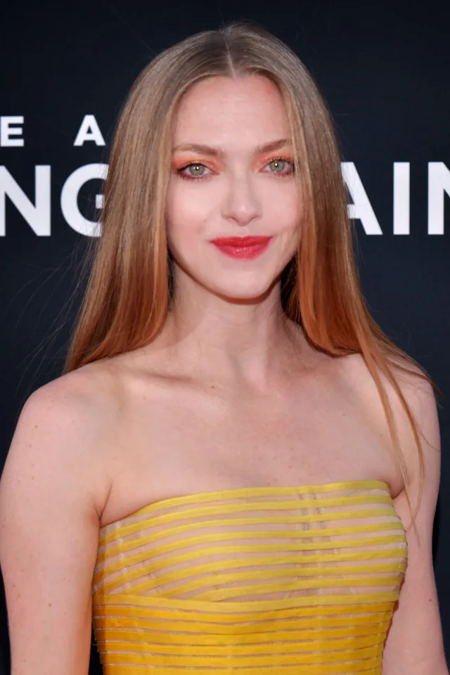 Amanda Seyfried smiling at the camera, with long straight hair and red lipstick, in a yellow striped strapless dress at an event.