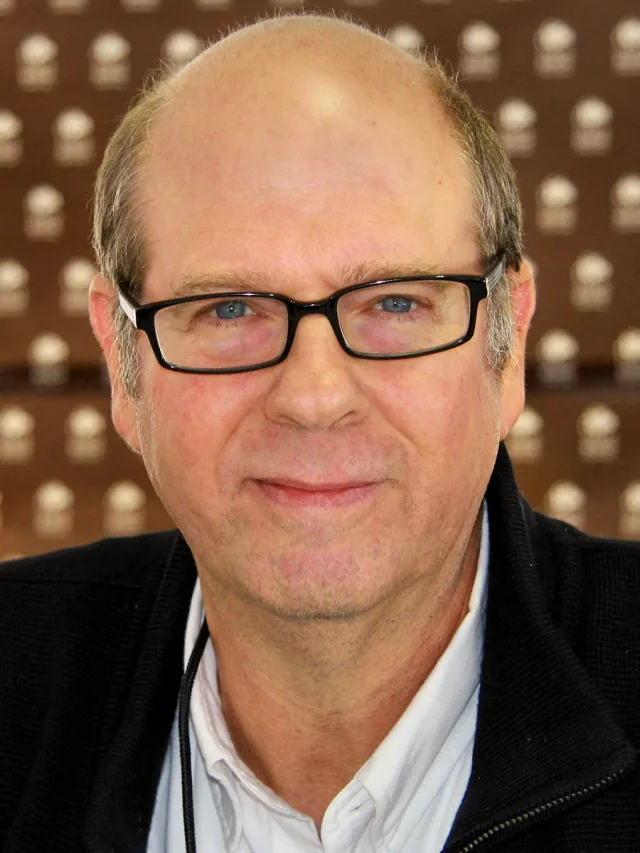 Close photograph of a balding man in black-framed glasses and a dark zip jacket over a white shirt, with a faint smile.