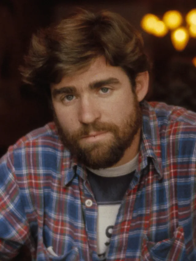 Young bearded man with tousled brown hair in a blue and red checked shirt, looking at the camera in a warmly lit room.