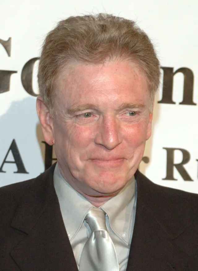 Close portrait of a smiling older man with short fair hair, wearing a dark jacket and a silver tie, in front of a white event backdrop.