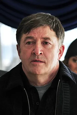 Portrait of a middle-aged man with short dark hair and a serious expression, wearing a dark zip-up jacket in front of a blue curtain.