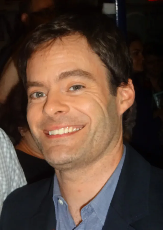 Close photograph of a smiling dark-haired man in a navy jacket and blue open-collar shirt, in a dim crowded room.
