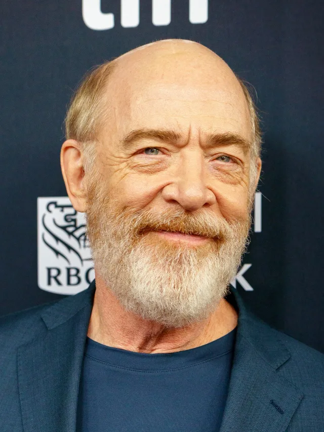 J. K. Simmons, bald with a white and ginger beard and a faint smile, wearing a navy jacket in front of a festival backdrop.