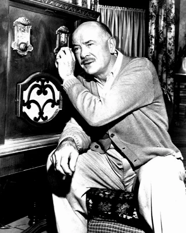 Black and white photograph of a bald, moustached man in a cardigan kneeling beside a large wooden radio cabinet with his hand raised to his head.