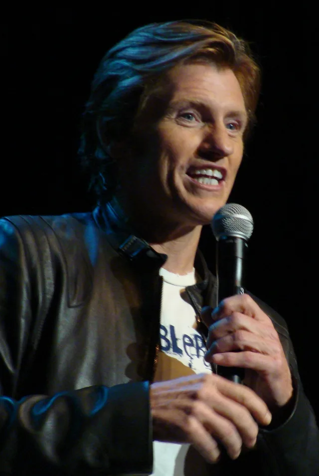 Denis Leary on stage holding a microphone, smiling, in a dark leather jacket against a black background.