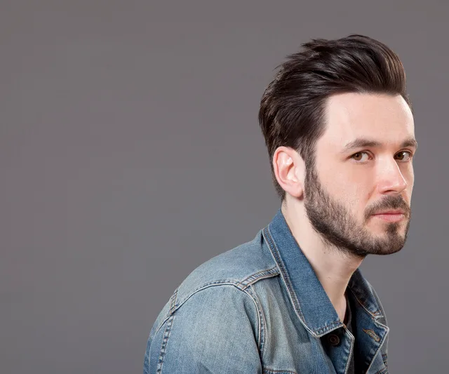 Man with a swept-back quiff and short beard, in a denim jacket, turned in profile against a plain grey backdrop.