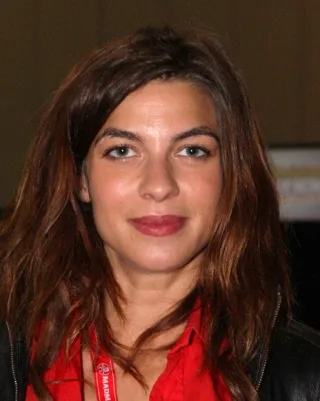 Portrait of a woman with long auburn hair and light eyes, wearing a red shirt and black leather jacket.