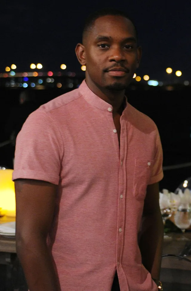 Aml Ameen in a pink short-sleeved shirt standing at an evening terrace with city lights blurred behind him.