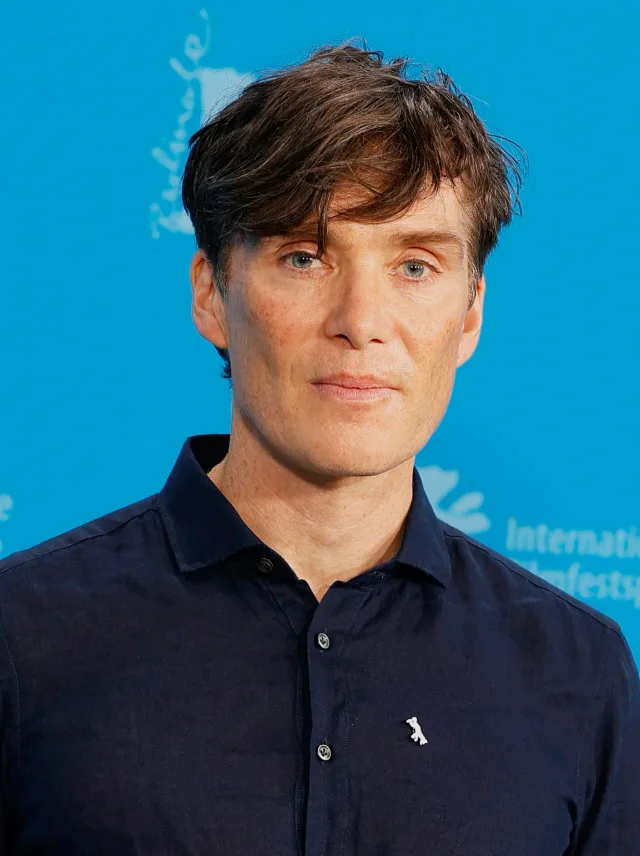 Head and shoulders photograph of Cillian Murphy with tousled brown hair, wearing a dark navy shirt against a blue backdrop.
