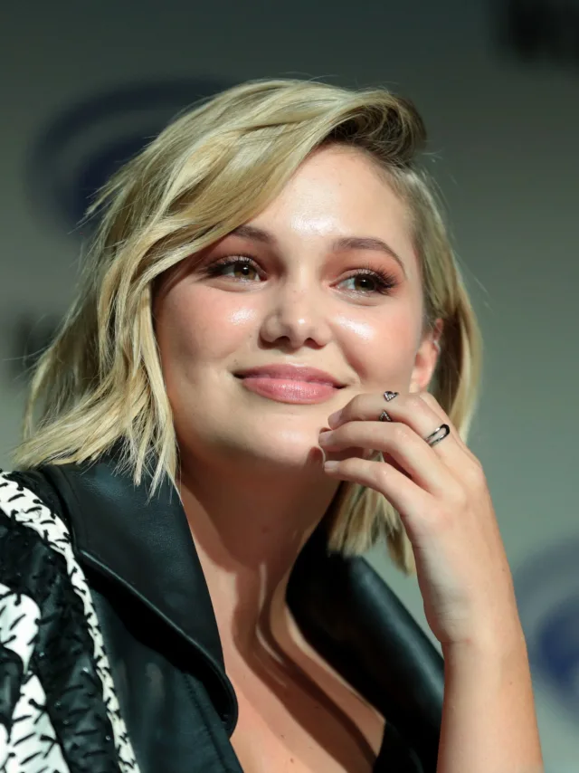 Close portrait of a smiling woman with a short blonde bob, resting her hand against her chin, wearing a black leather jacket at a panel event.