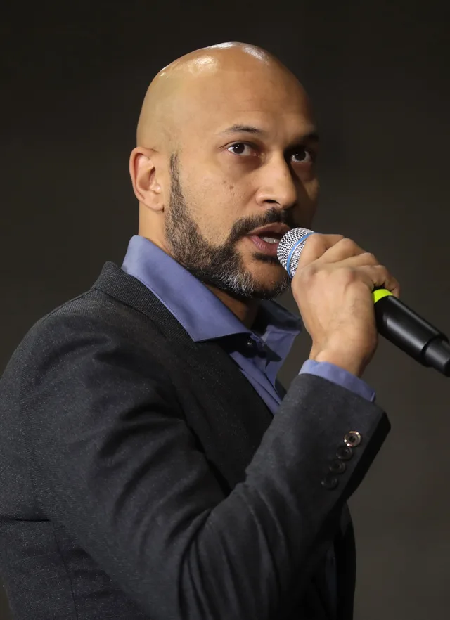 A bald man with a short beard in a dark jacket and blue shirt, speaking into a handheld microphone against a dark background.