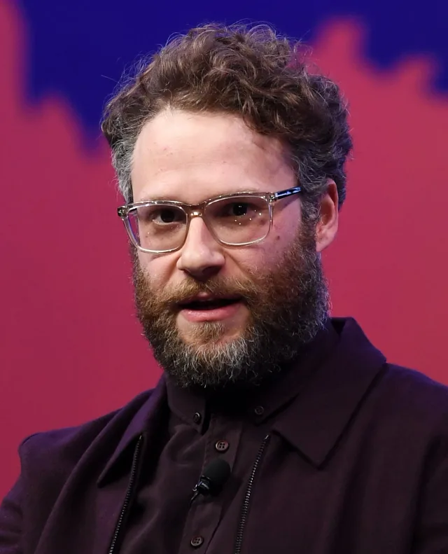 Close portrait of a bearded man with curly brown hair and clear-framed glasses, wearing a dark purple shirt, speaking against a red and blue backdrop.