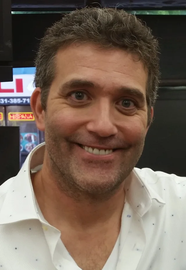 Close portrait of a smiling man with short dark, slightly greying hair and light stubble, wearing a white open-collared shirt.