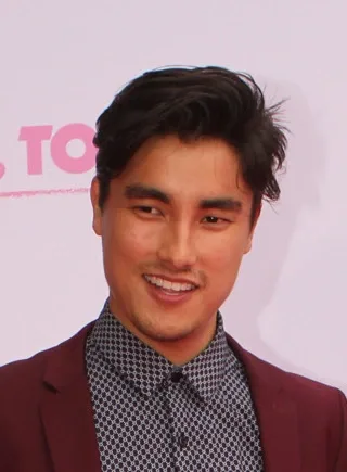 Head and shoulders photo of Remy Hii smiling, with dark swept hair, wearing a burgundy jacket over a patterned shirt.