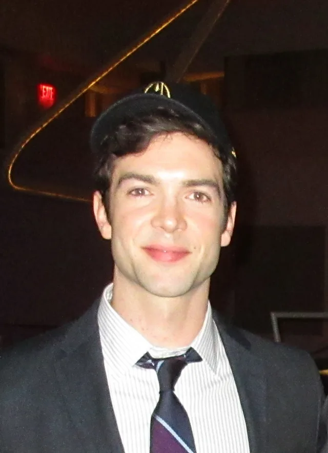 Head and shoulders photograph of a smiling man with dark hair, wearing a dark cap, a grey suit jacket, striped shirt and striped tie, indoors.
