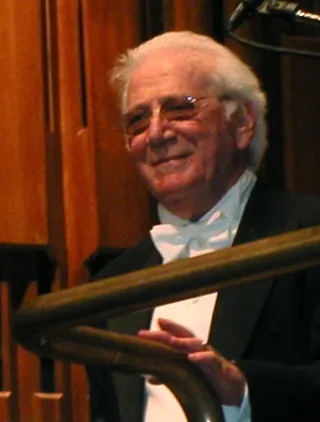 Elderly man with white hair and glasses, wearing a white bow tie and black jacket, smiling beside a wooden stair rail.