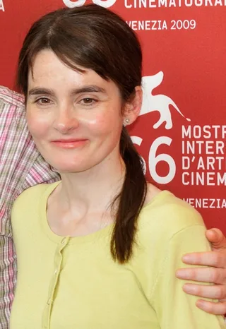 A woman with dark hair in a side ponytail and a pale green cardigan, smiling gently in front of a red festival backdrop.
