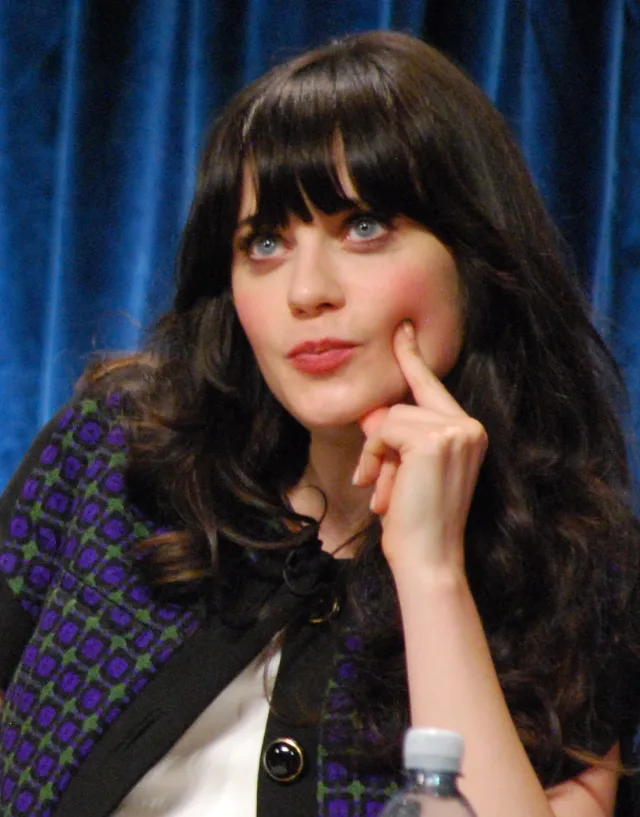 Zooey Deschanel with long dark hair and a fringe, resting a finger on her cheek and looking up, in front of a blue velvet curtain.