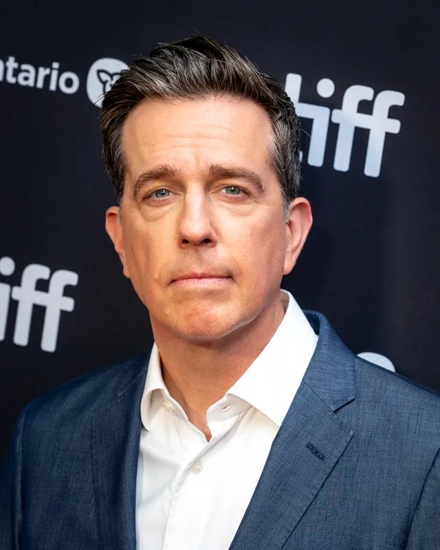 Head and shoulders photograph of a man with short dark swept hair, in a blue jacket and white open-necked shirt, in front of a festival backdrop.