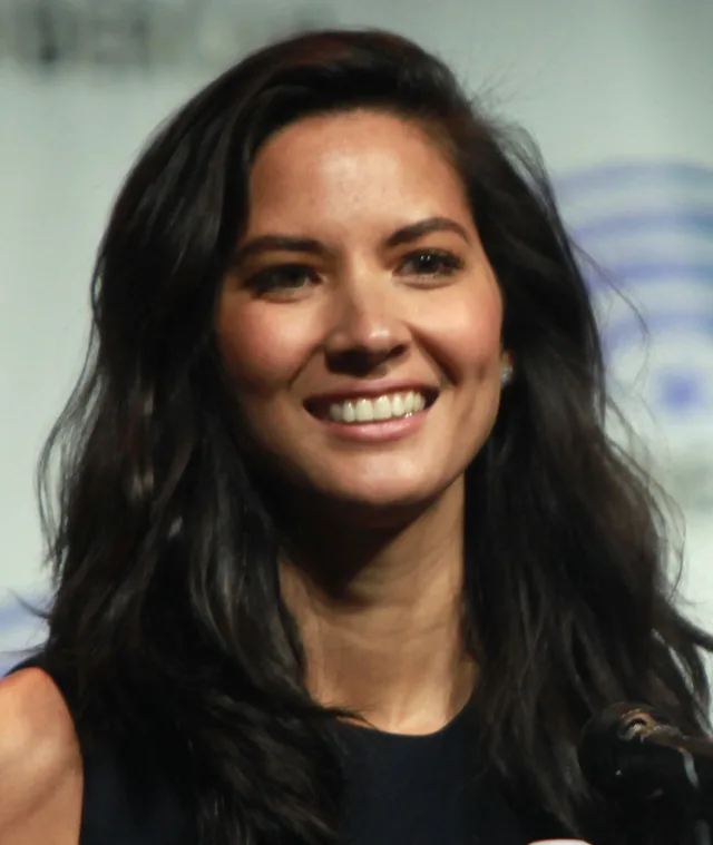 Portrait photograph of a smiling woman with long dark wavy hair, wearing a dark sleeveless top, with a microphone at the lower edge and a convention backdrop behind her.