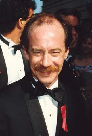 Smiling man with thinning reddish hair and a moustache, wearing a black bow tie and tuxedo with a red ribbon, outdoors in a crowd.
