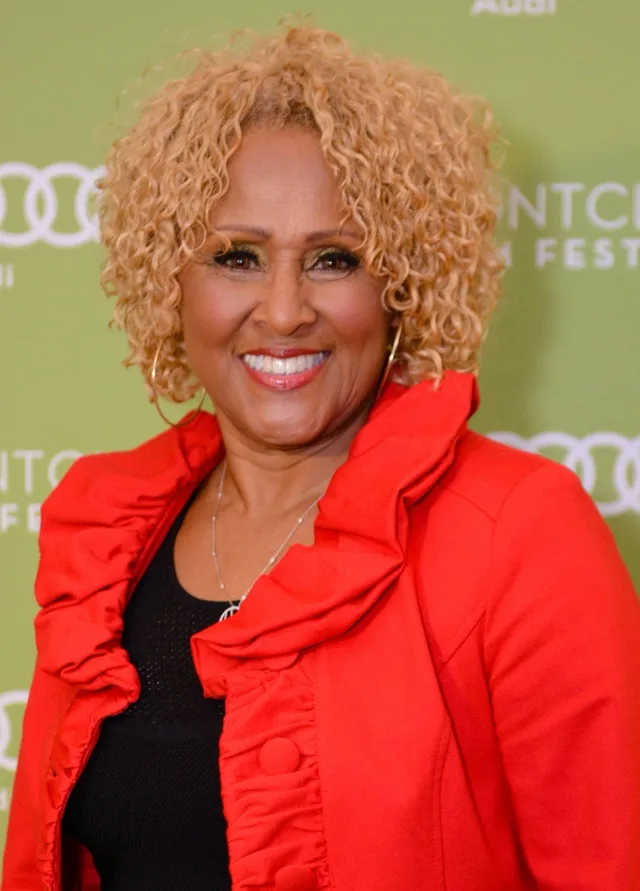 A smiling woman with short curly strawberry-blonde hair and gold hoop earrings, wearing a red ruffled jacket over black, in front of a green sponsor backdrop.