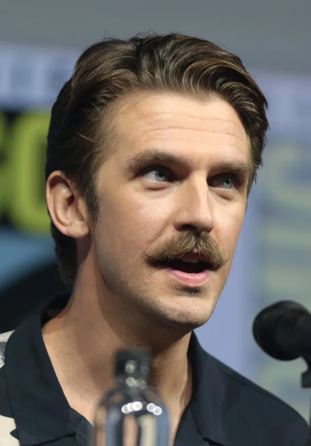 Close photograph of a man with swept-back brown hair and a moustache, speaking at a panel table with a microphone in front of him.
