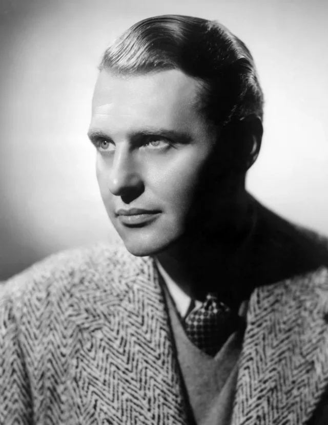Black and white studio portrait of a young man with slicked hair in a herringbone coat, looking off to the side.