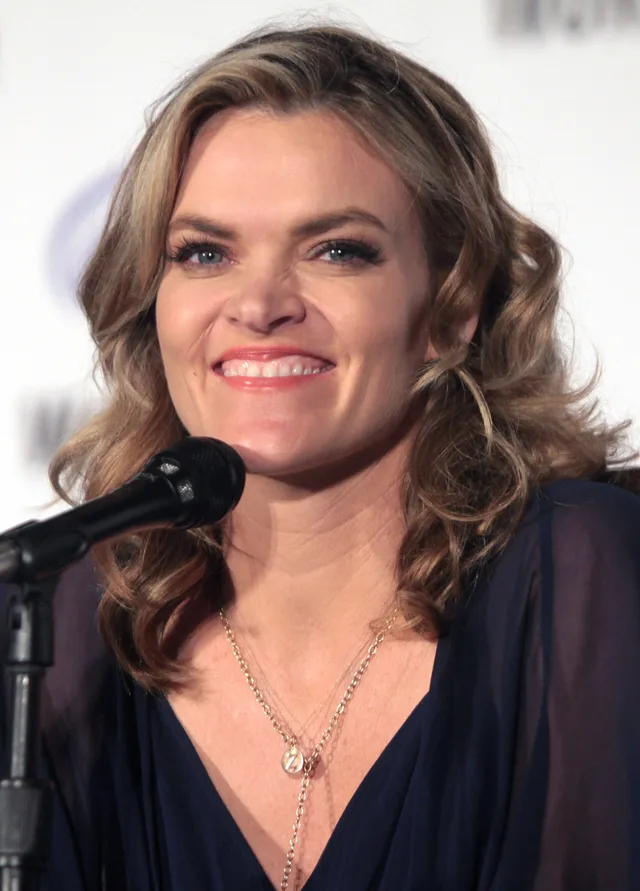 A smiling woman with wavy blonde hair and a navy top, seated at a microphone at a convention panel.