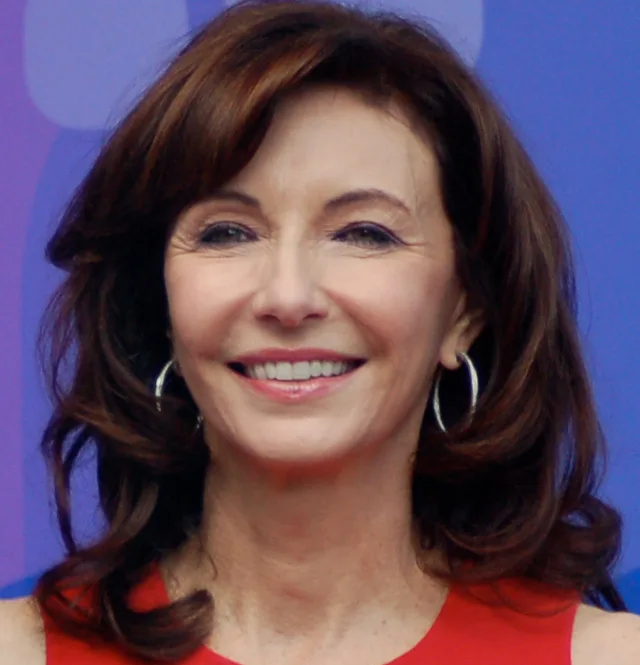 A smiling woman with shoulder-length auburn hair and hoop earrings, wearing a red top, photographed against a purple and blue backdrop.
