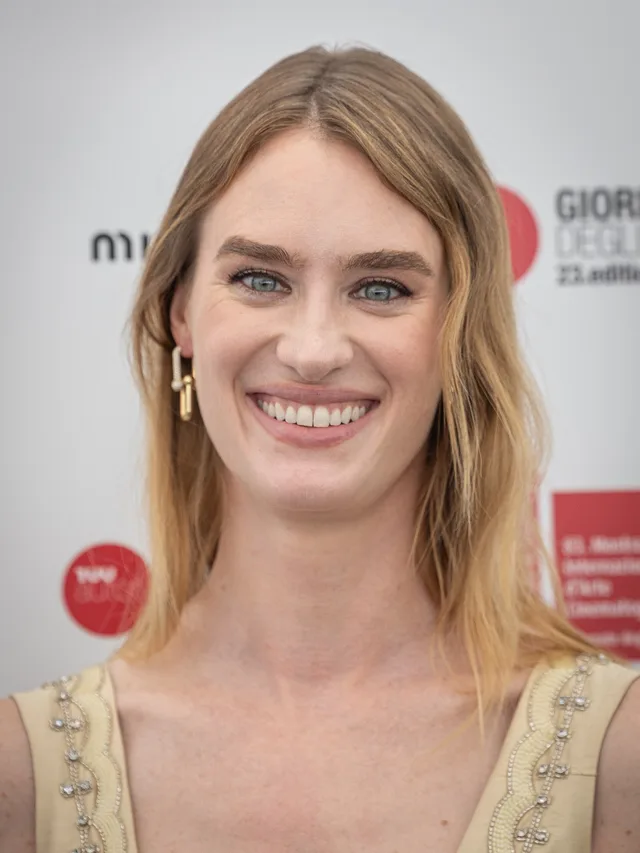 A smiling woman with straight, light brown hair and blue eyes, wearing a cream top with beaded trim and a gold earring, in front of a festival backdrop.