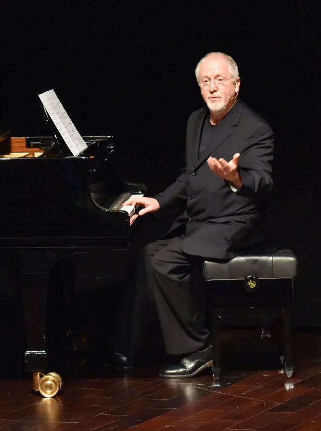 A white-haired, bearded man in glasses and a dark jacket sits at a grand piano on a dark stage, one hand on the keys, talking to an audience.