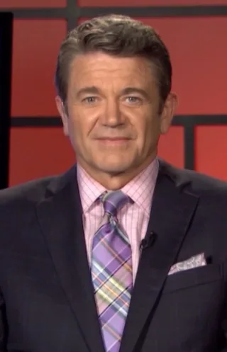 Smiling man with short dark hair and blue eyes in a dark suit, pink checked shirt and purple plaid tie, in front of a red studio backdrop.